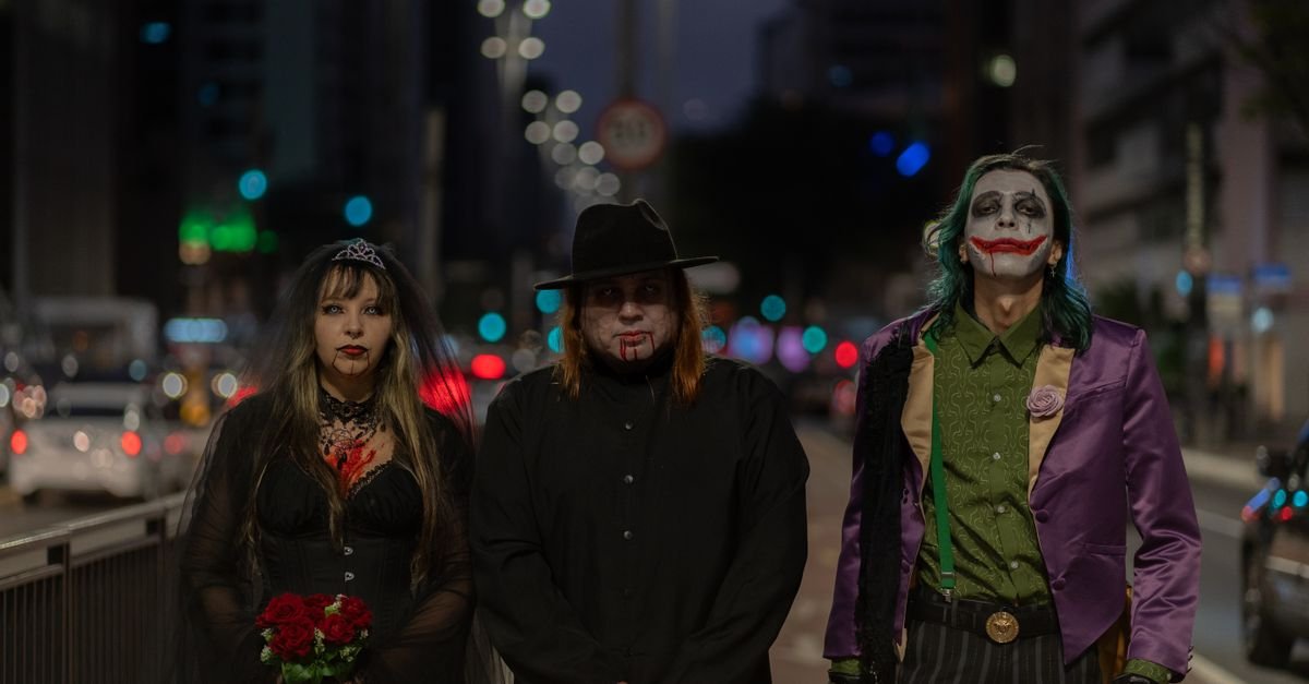Três pessoas fantasiadas de personagens góticos e de palhaço posam em rua iluminada à noite