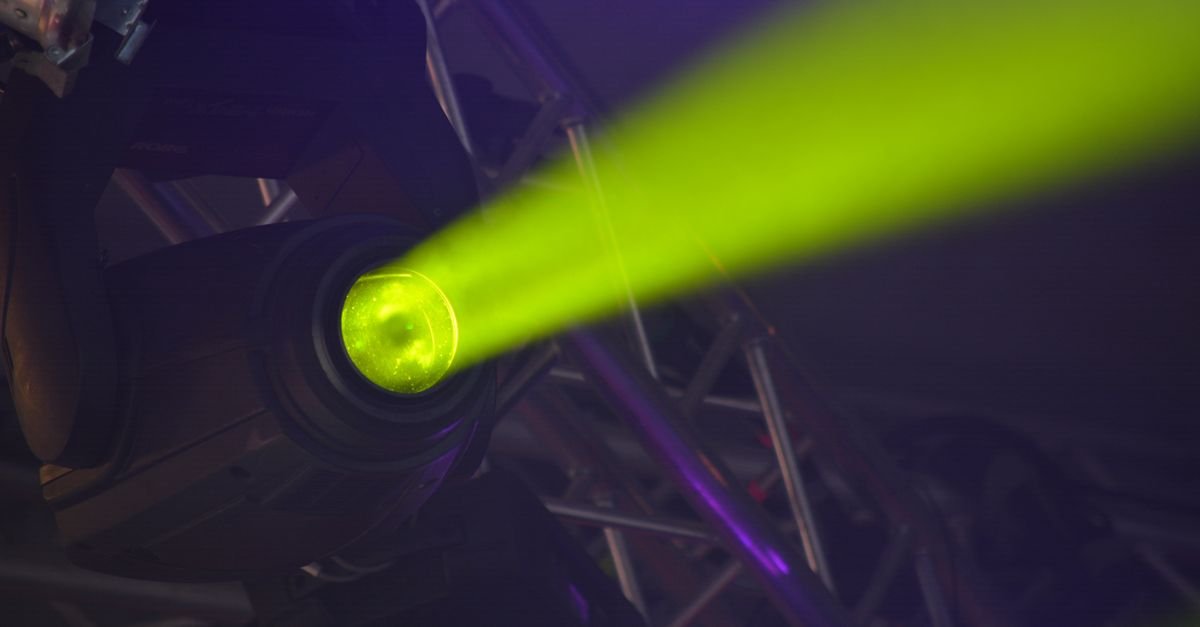 Refletor de palco aceso projetando feixe de luz verde sobre estrutura metálica de iluminação