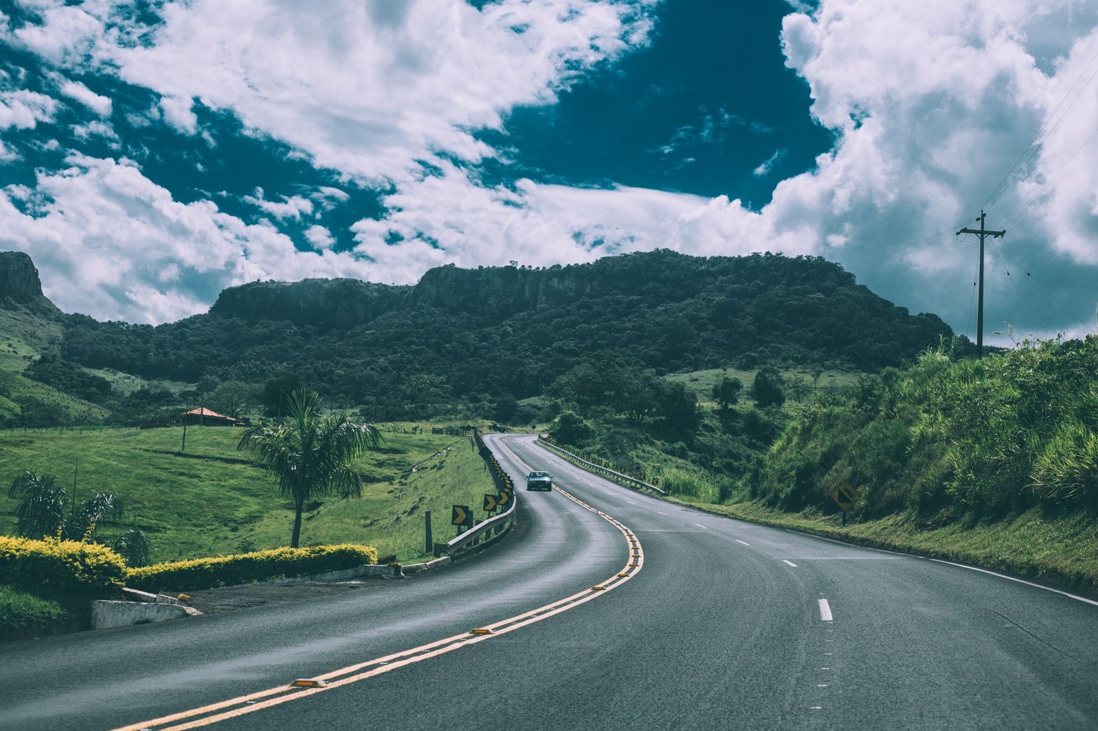 Rodovia de pista simples com faixa continua amarela dupla no centro, curva a frente e um carro ao fundo