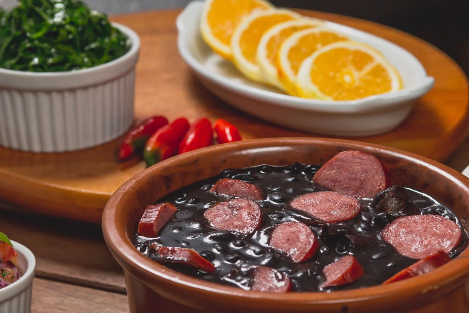 Panela de barro com feijoada, linguiça fatiada, couve e laranja ao fundo