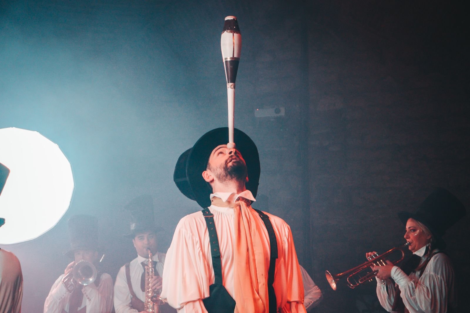 Artista equilibra um pino de malabarismo no nariz diante de músicos de cartola e figurino de época
