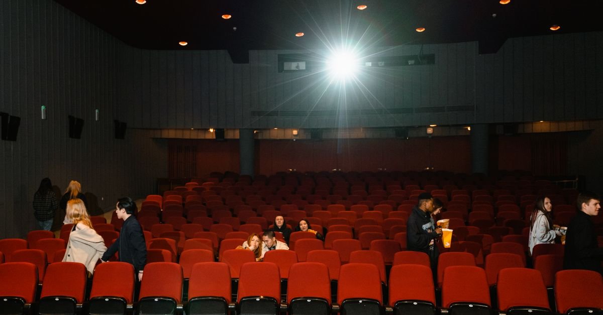 Sala de cinema com poltronas vermelhas e público entrando antes da sessão