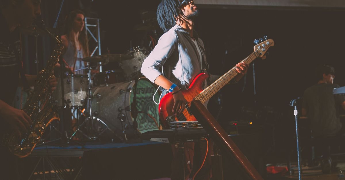 Banda tocando ao vivo em palco, com baixista em primeiro plano e sopro ao fundo