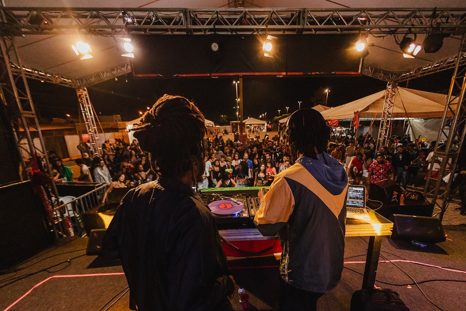 Dois DJs de costas comandam a discotecagem em palco ao ar livre diante do público, à noite