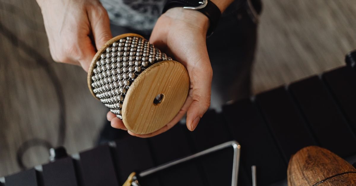 Maos segurando uma cabasa, instrumento de percussao, sobre uma mesa com outros instrumentos musicais