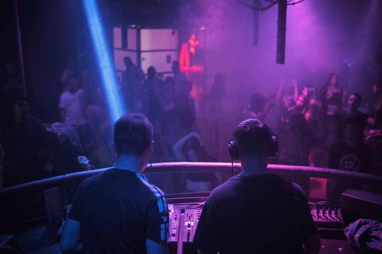 Dois DJs de costas diante da pista de uma casa noturna iluminada em roxo, foto ilustrativa