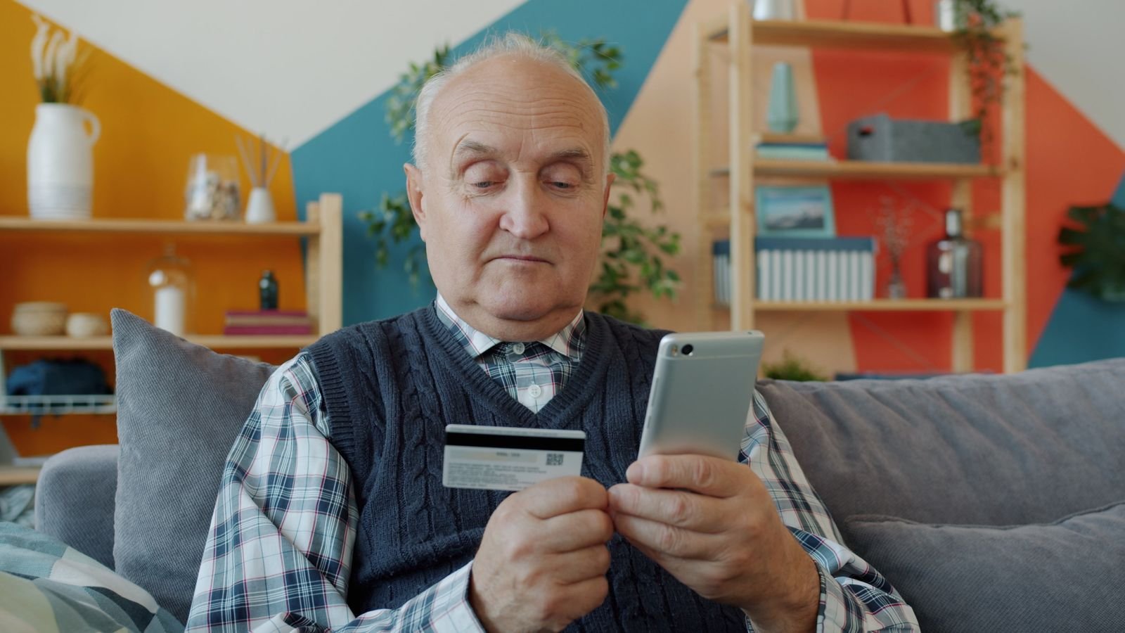 Homem idoso sentado no sofá segura um cartão de crédito e um celular, imagem de arquivo