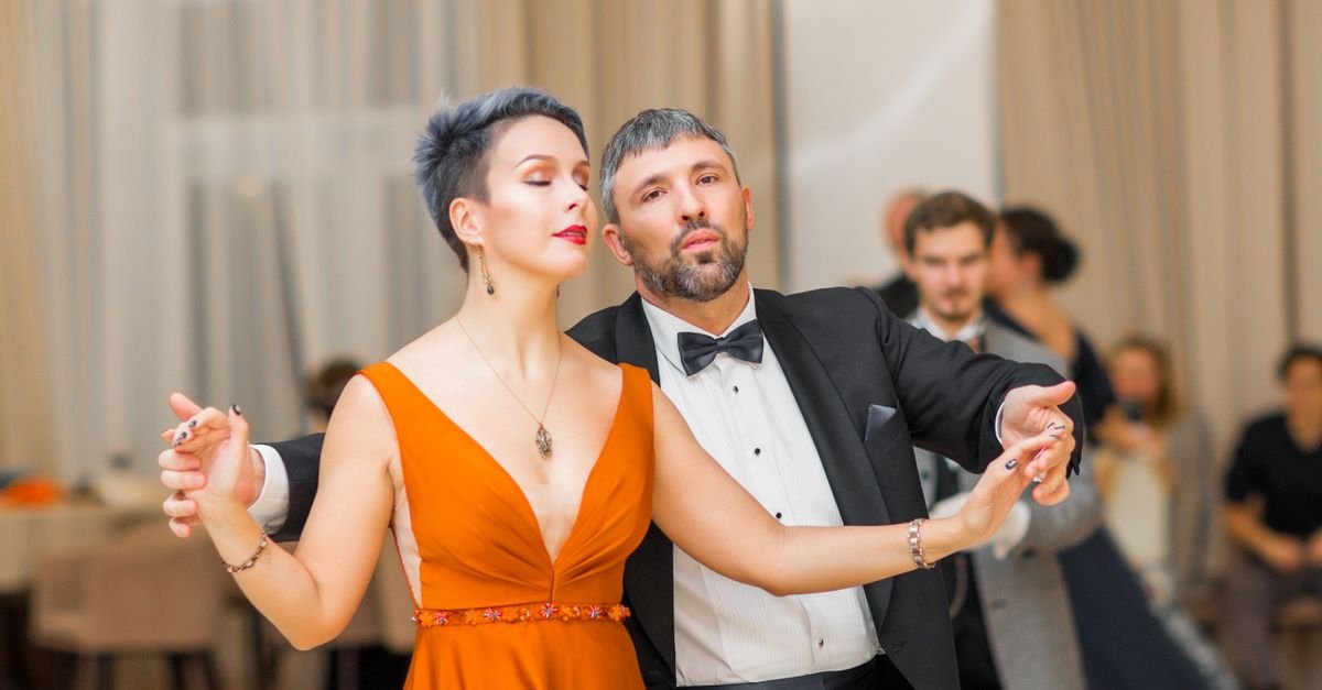 Casal dançando em salão, ela de vestido laranja e ele de terno com gravata-borboleta