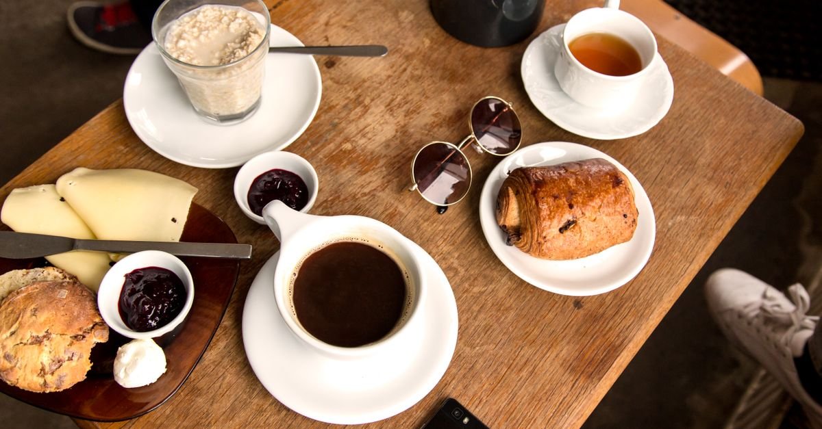 Mesa de madeira em cafeteria com xícara de café, pão, queijo, geleia e croissant