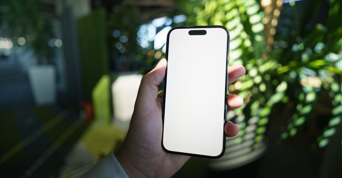 Mao segurando um telefone celular com a tela em branco, em ambiente com plantas ao fundo