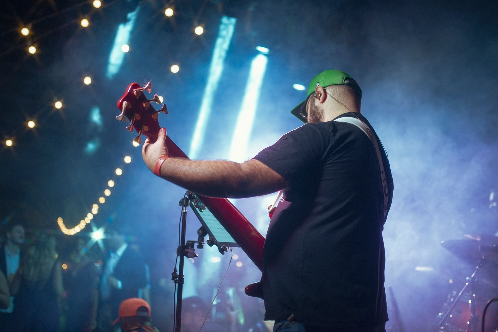Baixista visto de costas em palco enfumaçado, com luzes azuis e cordão de lâmpadas ao fundo