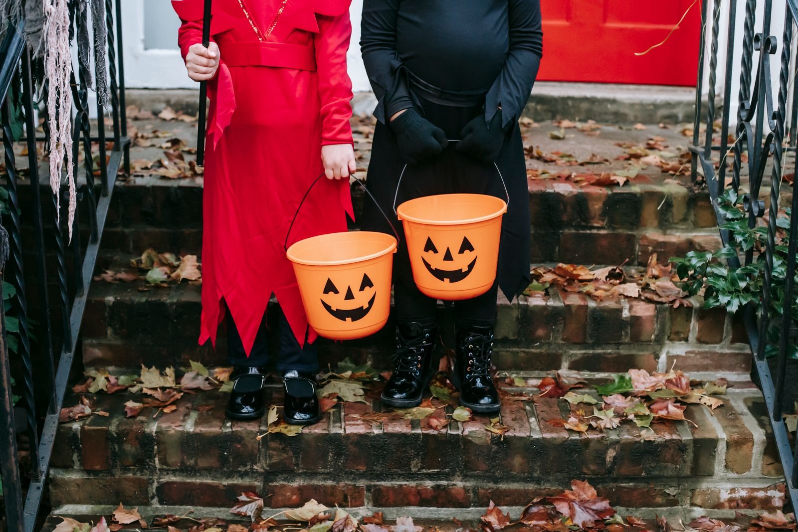 Duas crianças fantasiadas seguram baldes laranja em forma de abóbora de Halloween numa escada de tijolos; imagem ilustrativa de banco de fotos