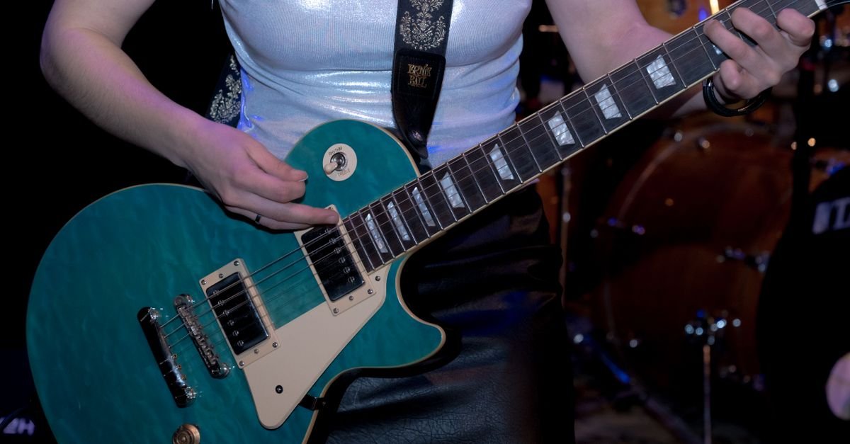 Guitarra elétrica verde-azulada sendo tocada em palco, com bateria desfocada ao fundo