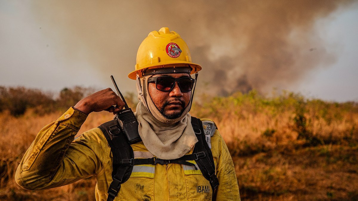 Brigadista do Ibama com rádio em campo de vegetação seca e fumaça ao fundo