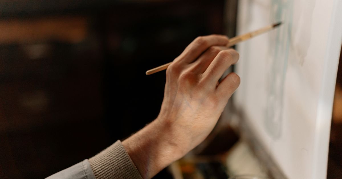 Mão de artista segurando pincel enquanto pinta uma tela em ateliê