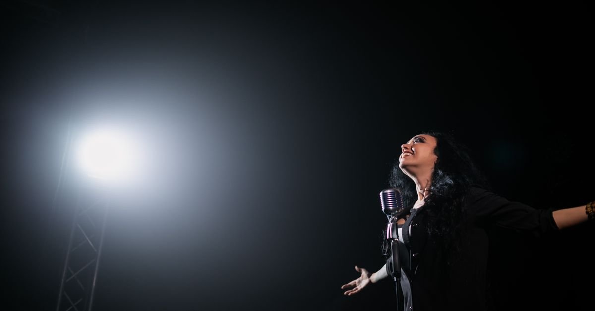 Cantora se apresenta ao microfone em palco escuro iluminado por um refletor