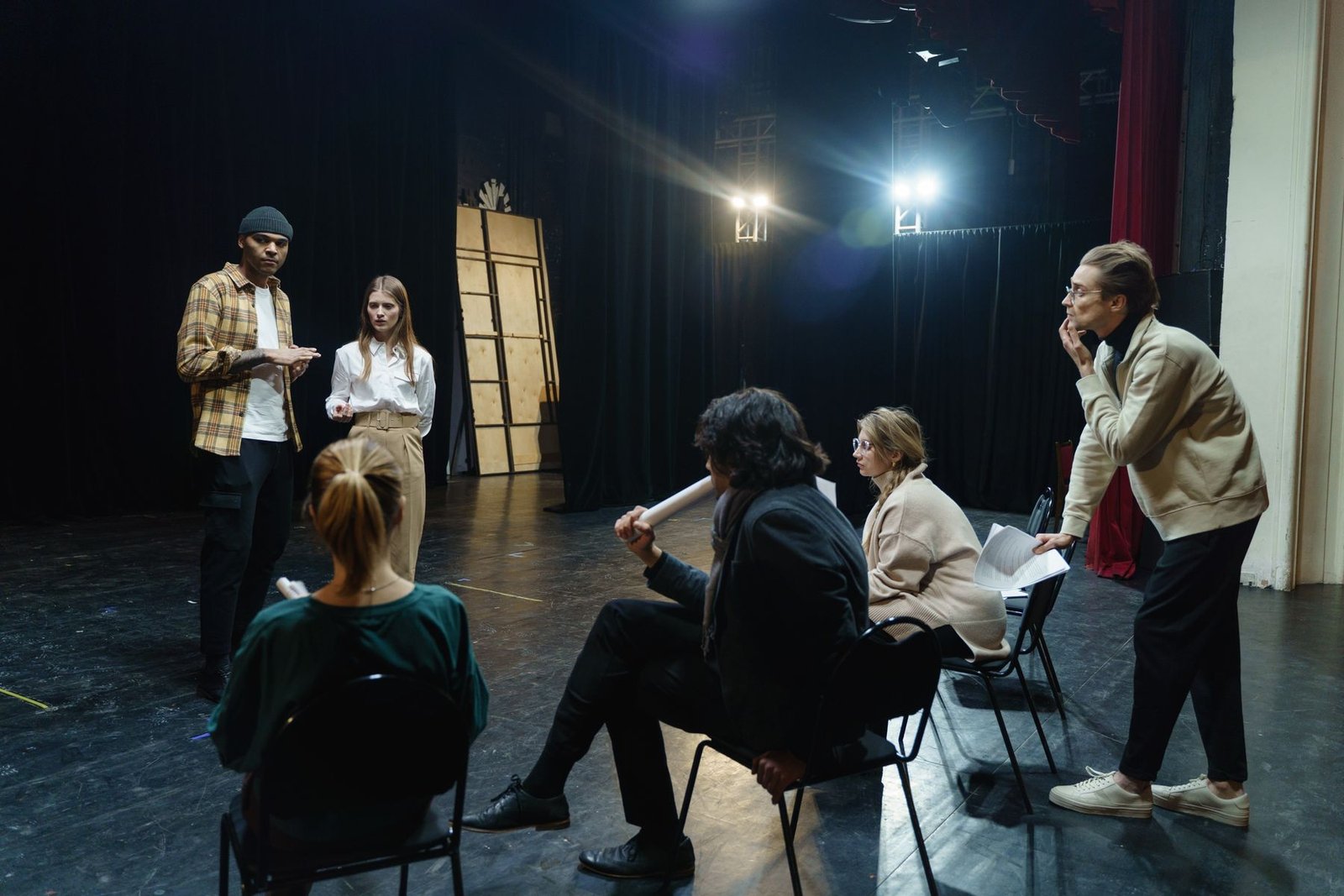 Elenco e diretor reunidos no palco durante um ensaio, com refletores acesos