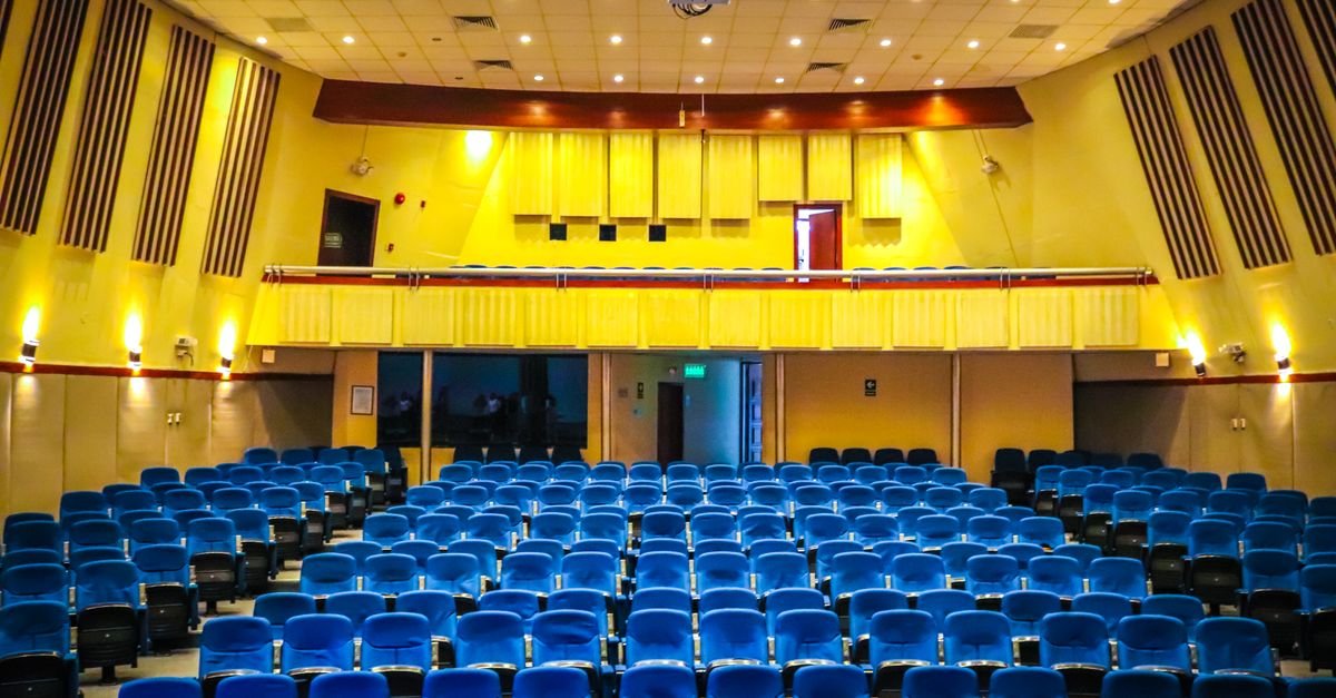 Auditório com poltronas azuis e paredes amarelas visto do fundo da plateia