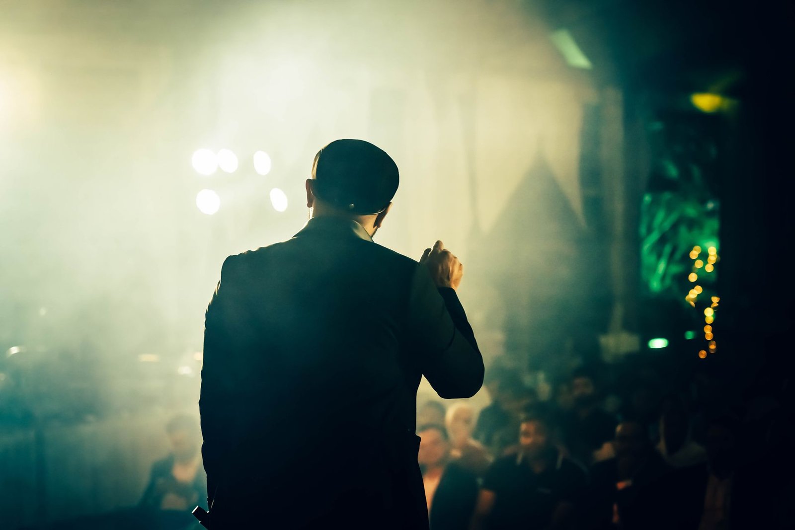 Humorista de costas no palco, diante da plateia, sob luzes de show