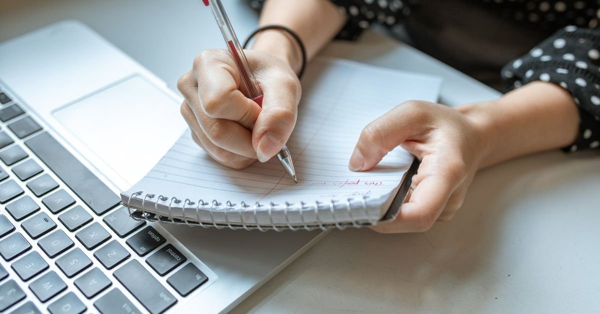 Maos escrevendo em caderno espiral ao lado de notebook sobre mesa