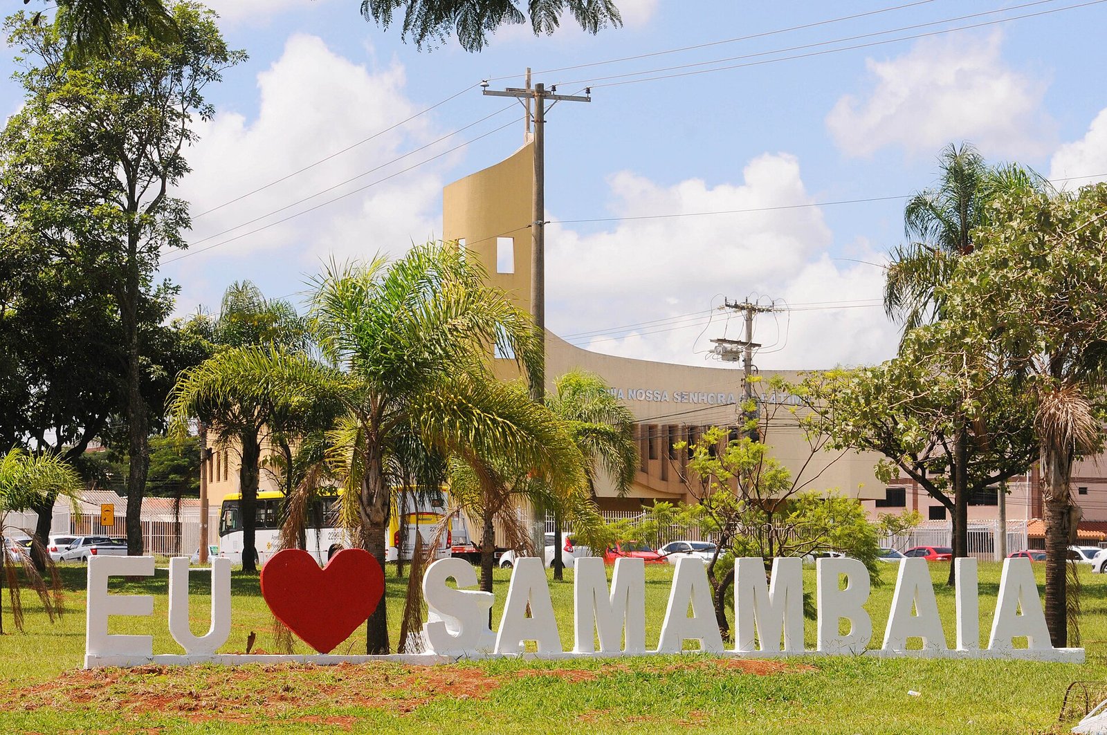 Letreiro com os dizeres Eu amo Samambaia em gramado, com igreja e palmeiras ao fundo