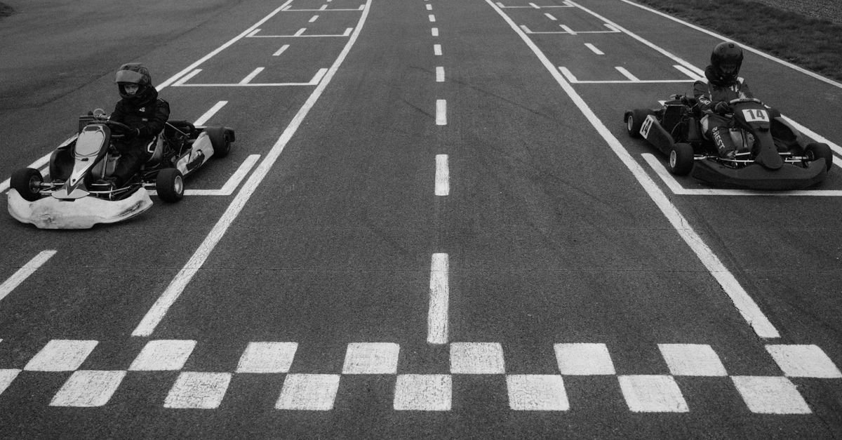 Dois karts alinhados na linha de largada de uma pista de kart, com pilotos de capacete