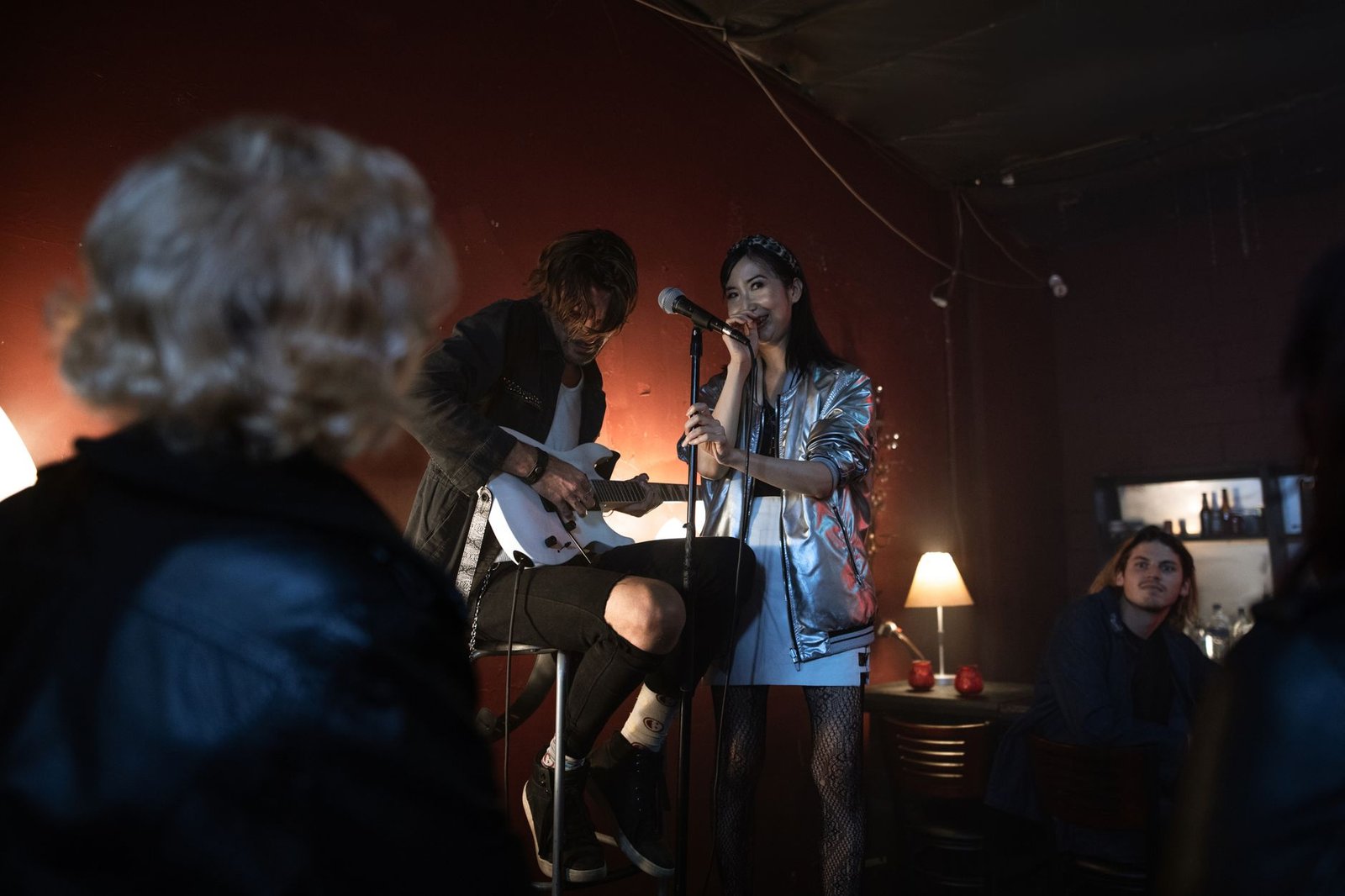 Cantora ao microfone e guitarrista sentado em um palco pequeno de bar, com plateia em primeiro plano; foto ilustrativa de show intimista