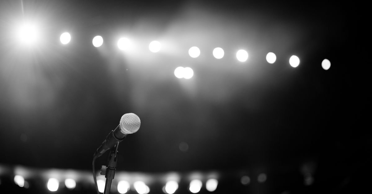 Microfone em pedestal no palco, com refletores acesos ao fundo, em foto em preto e branco