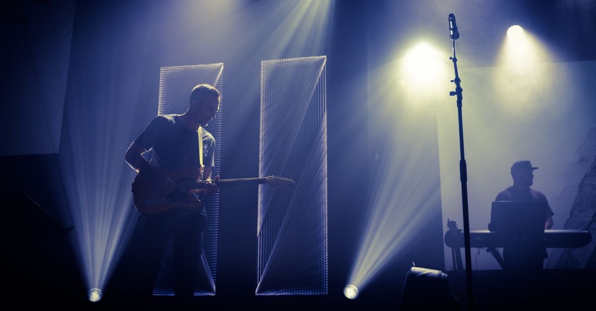 Músico toca guitarra em contraluz no palco, com DJ ao fundo e feixes de luz