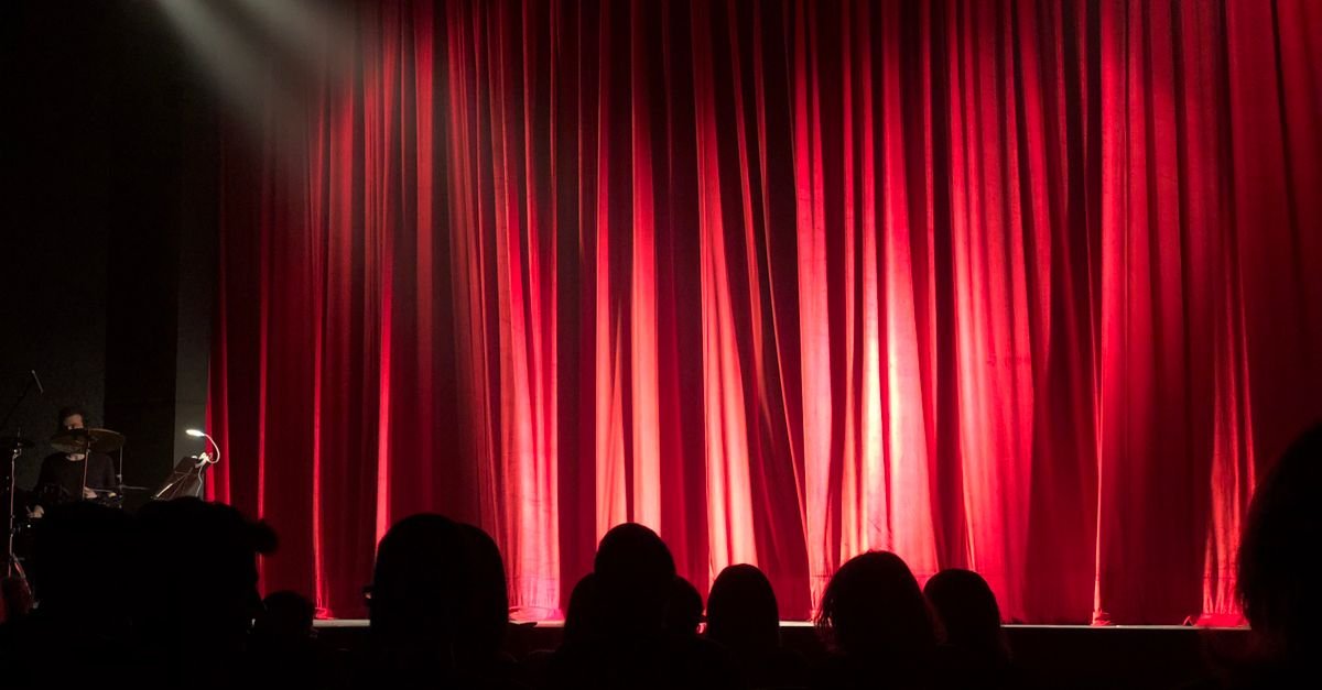 Cortina vermelha de teatro iluminada por refletor, com plateia em silhueta