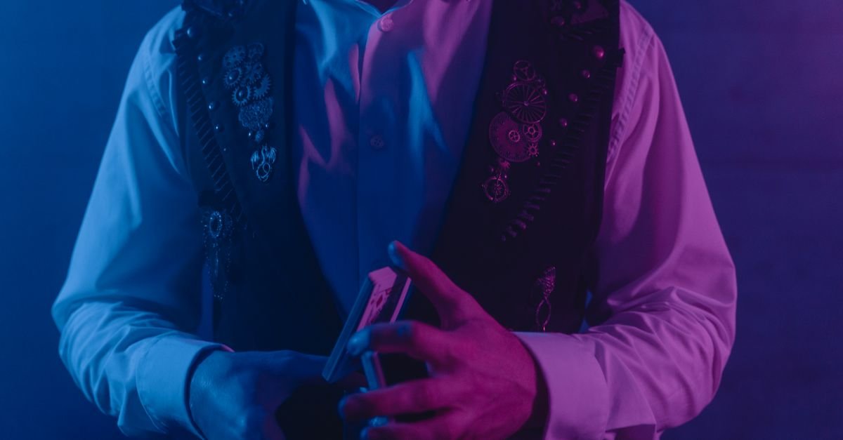 Magico de colete manipula baralho sob luz azul e roxa em palco