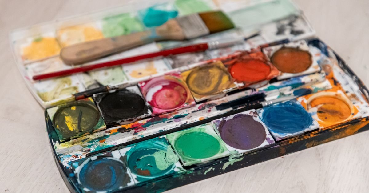 Paleta de aquarela usada, com tintas coloridas e pinceis apoiados sobre a caixa