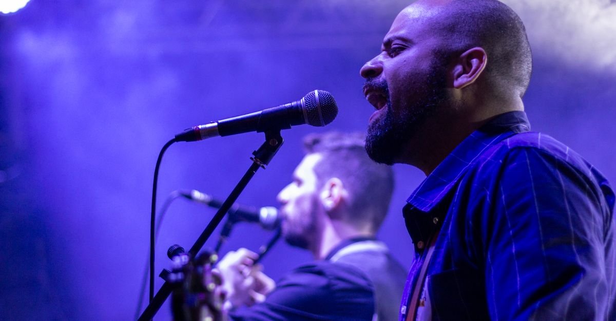 Cantor ao microfone durante show, com iluminação roxa e violonista ao fundo