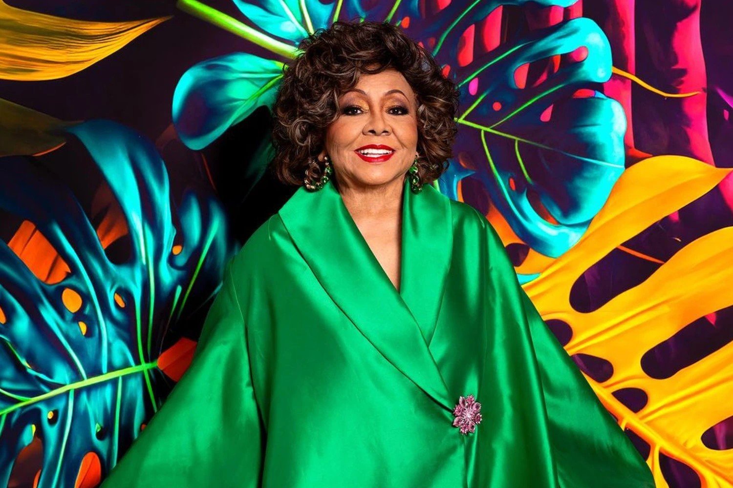 A cantora Alcione sorri em frente a um painel colorido, vestindo um manto verde