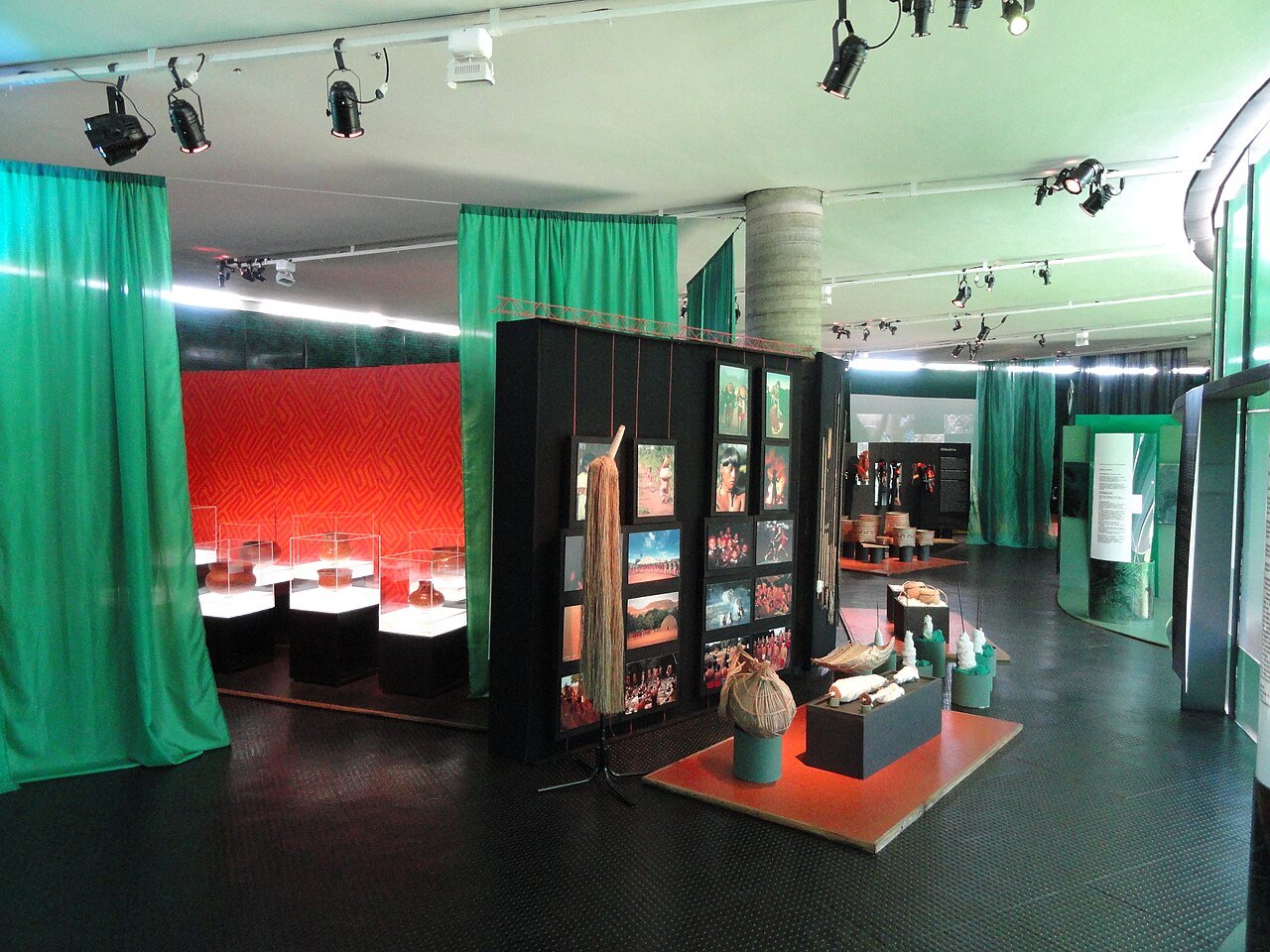 Sala de exposição do Memorial dos Povos Indígenas com cortinas verdes, vitrines de cerâmica e painéis de fotografias
