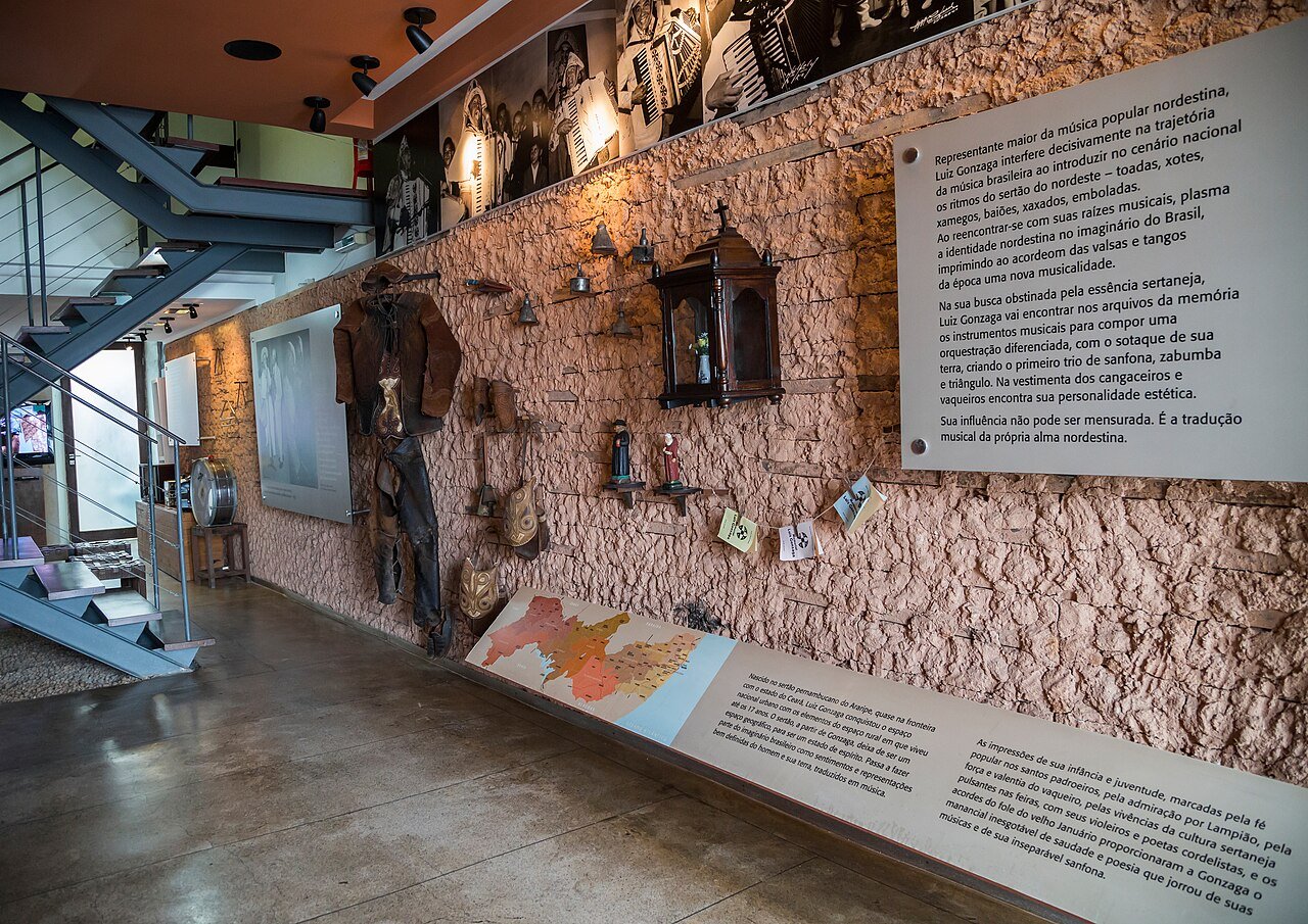 Sala do Memorial Luiz Gonzaga, no Recife, com figurino de vaqueiro, fotos e painéis na parede de pedra