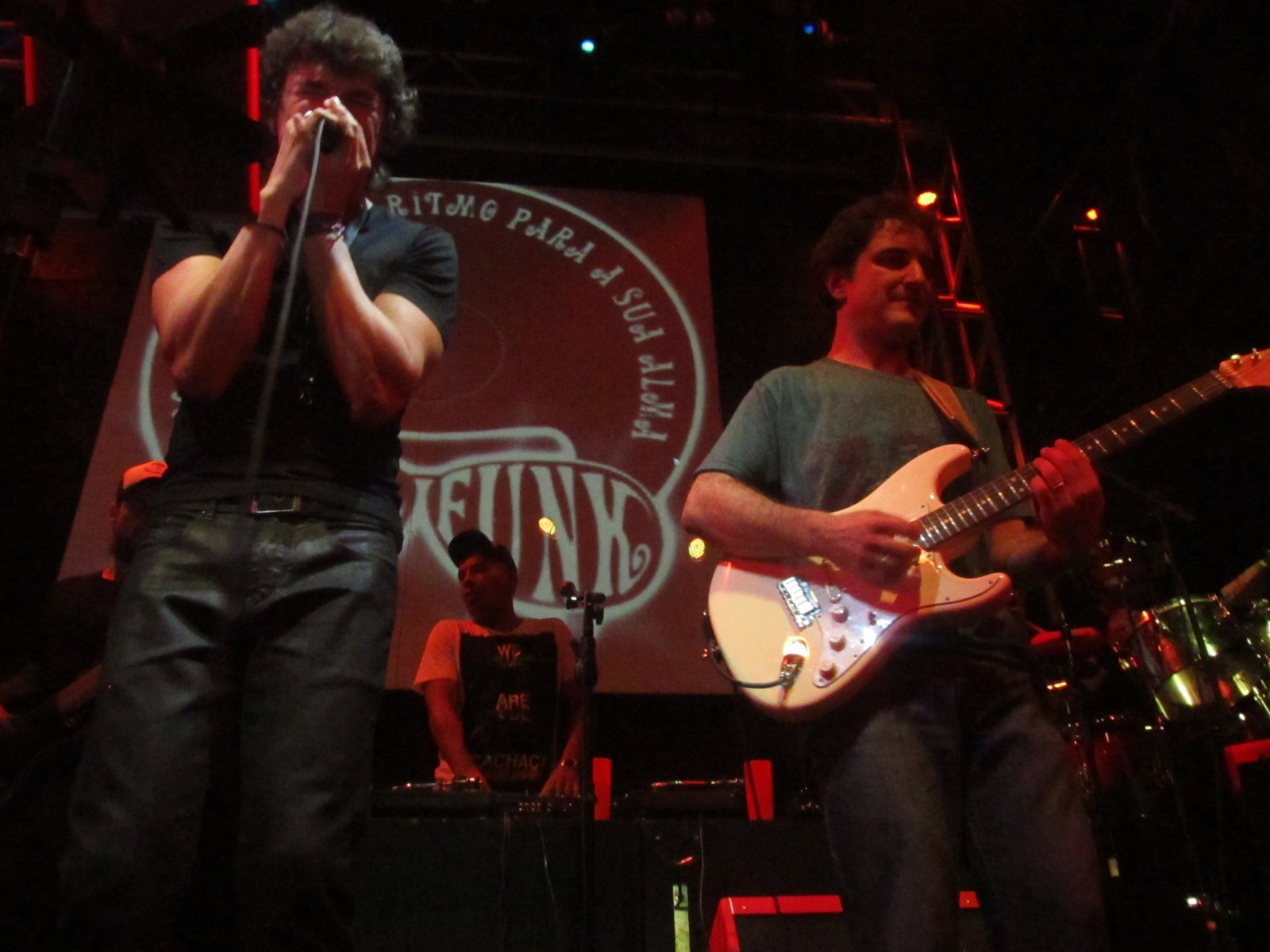Milton Guedes tocando harmônica em show com a banda Soul Funk, com guitarrista ao lado e telão vermelho ao fundo