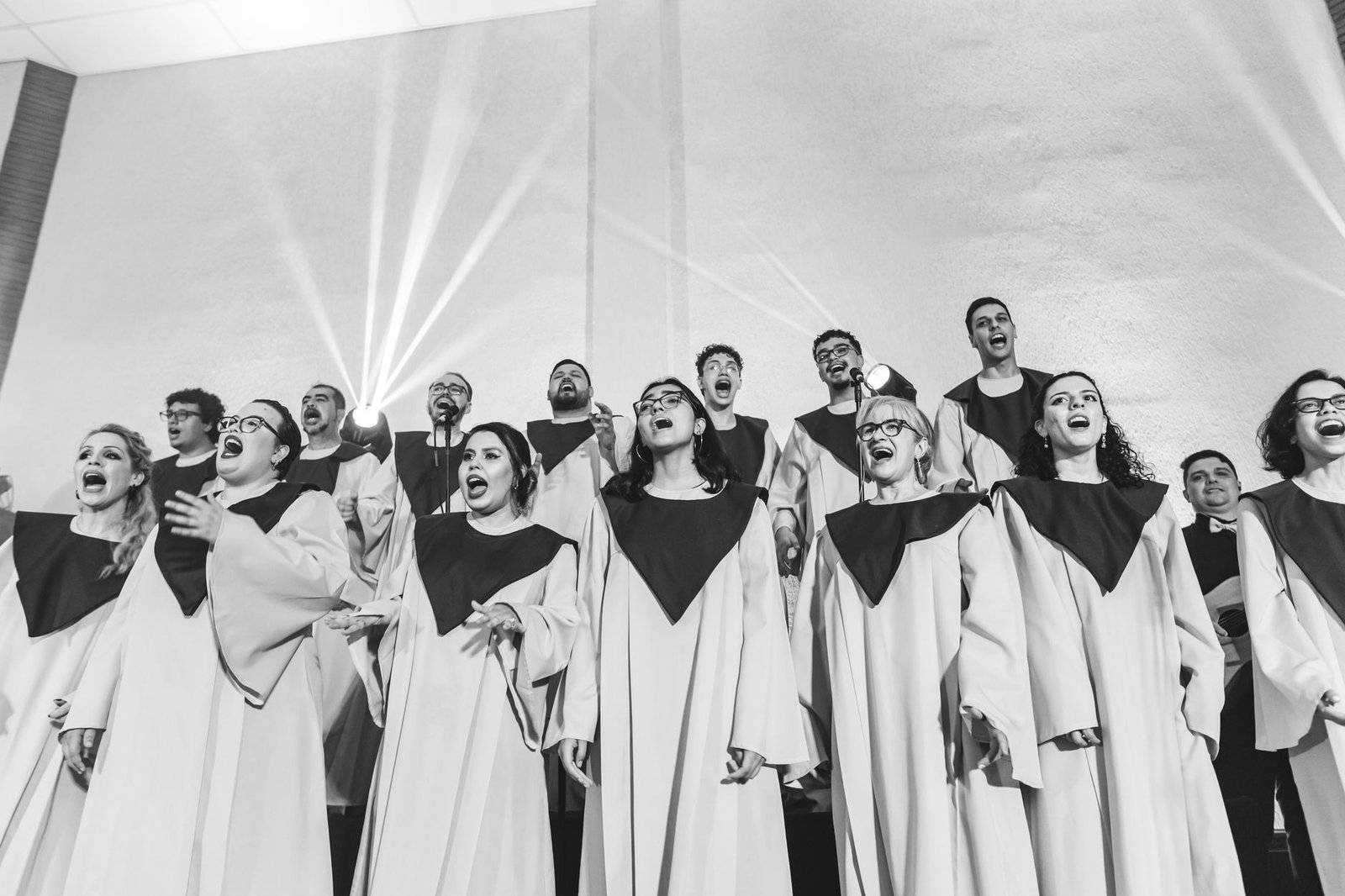 Coral de vozes femininas e masculinas em vestes claras com gola escura, cantando em palco com feixes de luz, em foto em preto e branco