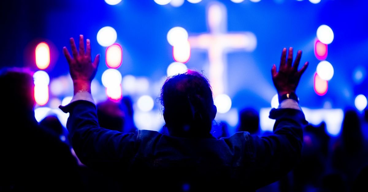 Pessoa de braços erguidos diante de palco iluminado de azul com uma cruz ao fundo