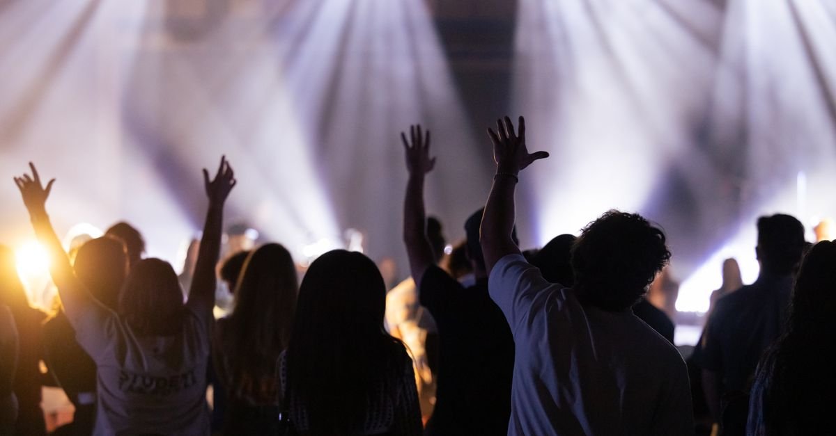 Plateia em silhueta com mãos erguidas diante de palco iluminado em show de música cristã