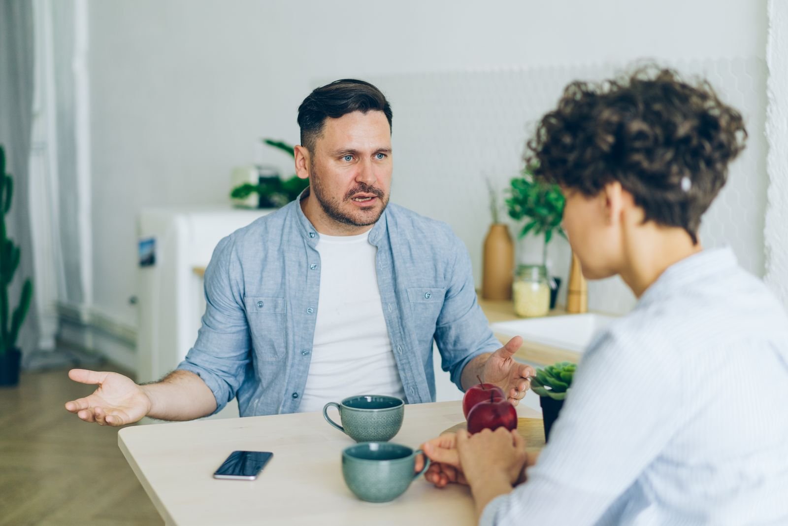 Duas pessoas conversam à mesa de uma cozinha, com xícaras e um celular, imagem de arquivo