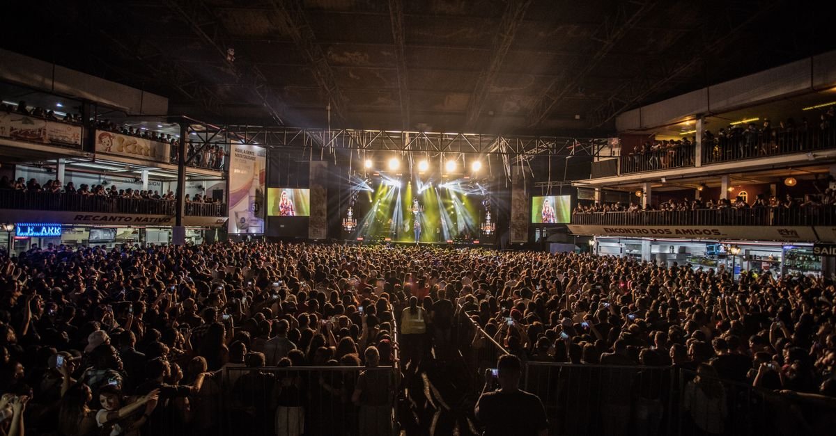 Público lotando pavilhão durante show com palco iluminado ao fundo