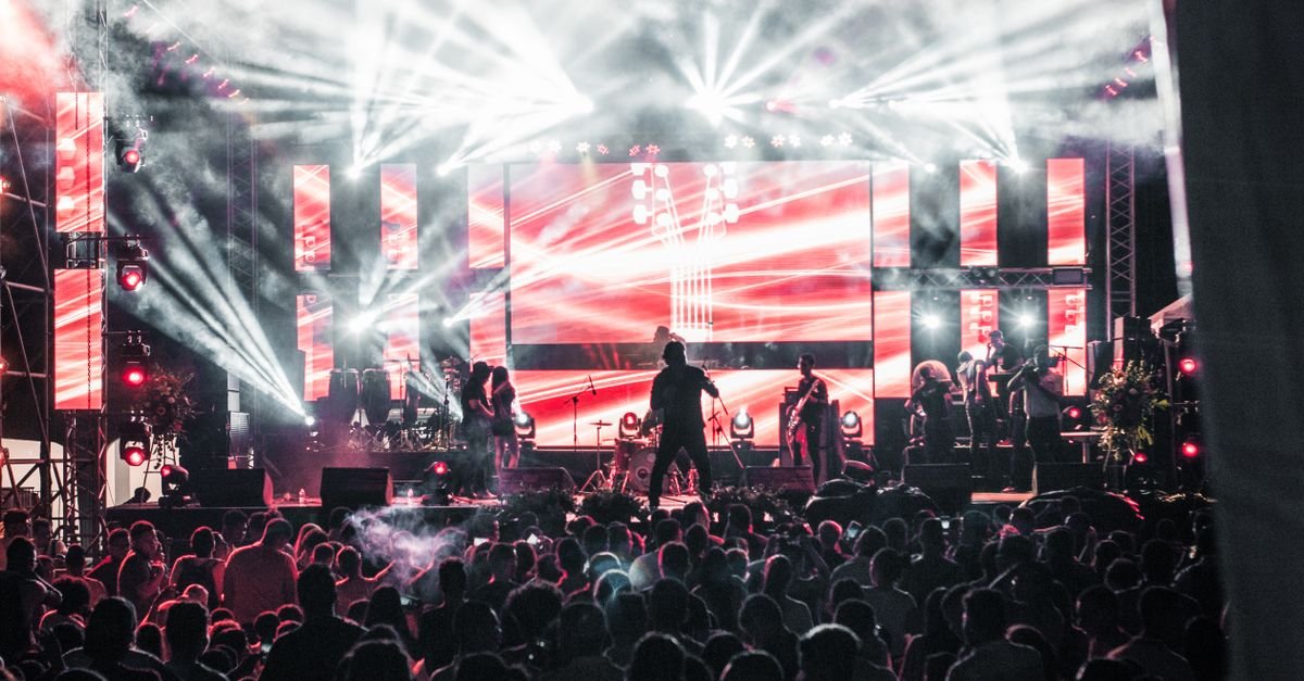 Palco de festival iluminado à noite com público em silhueta diante do telão