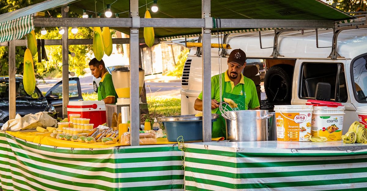 Barraca de comida em feira ao ar livre, com toldo listrado e vendedores preparando pratos