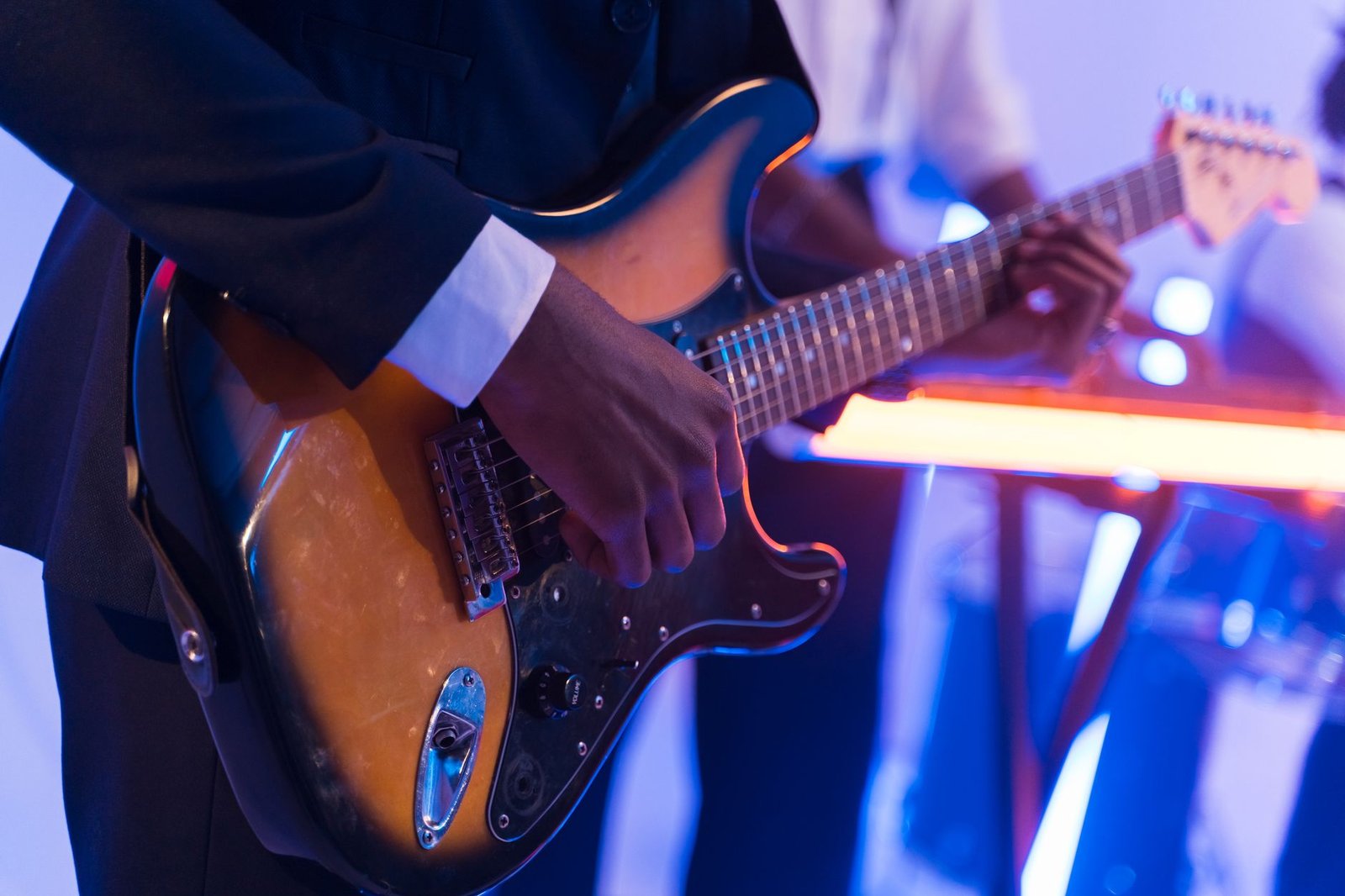 Mãos de um guitarrista de terno tocando uma guitarra elétrica sob luz azul e laranja