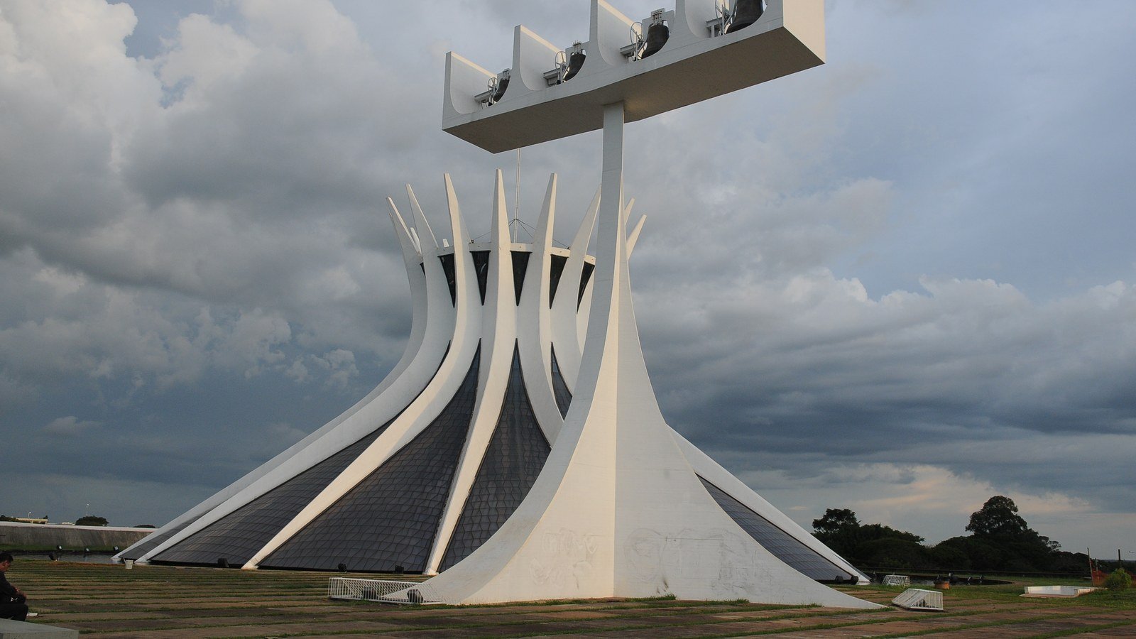 Fachada da Catedral Metropolitana de Brasília com o campanário de quatro sinos em primeiro plano, sob céu nublado