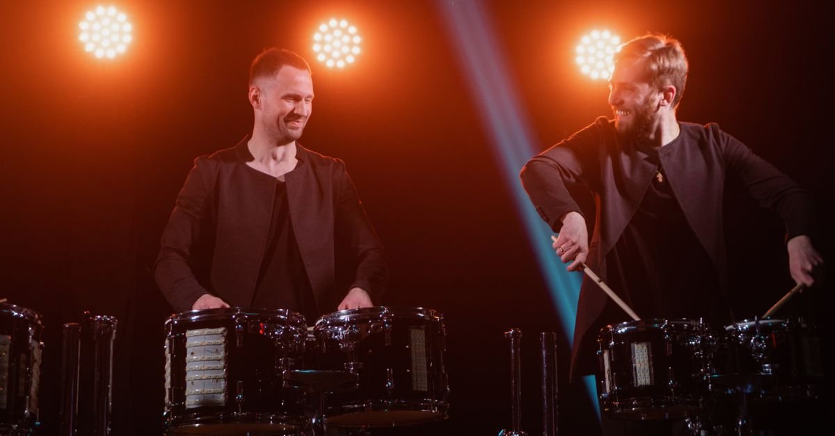 Dois percussionistas tocando tambores sob luzes de palco em apresentação ao vivo