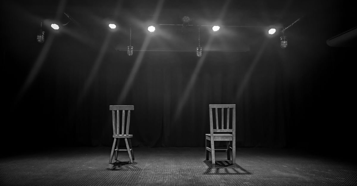 Palco de teatro vazio, em preto e branco, com duas cadeiras sob refletores