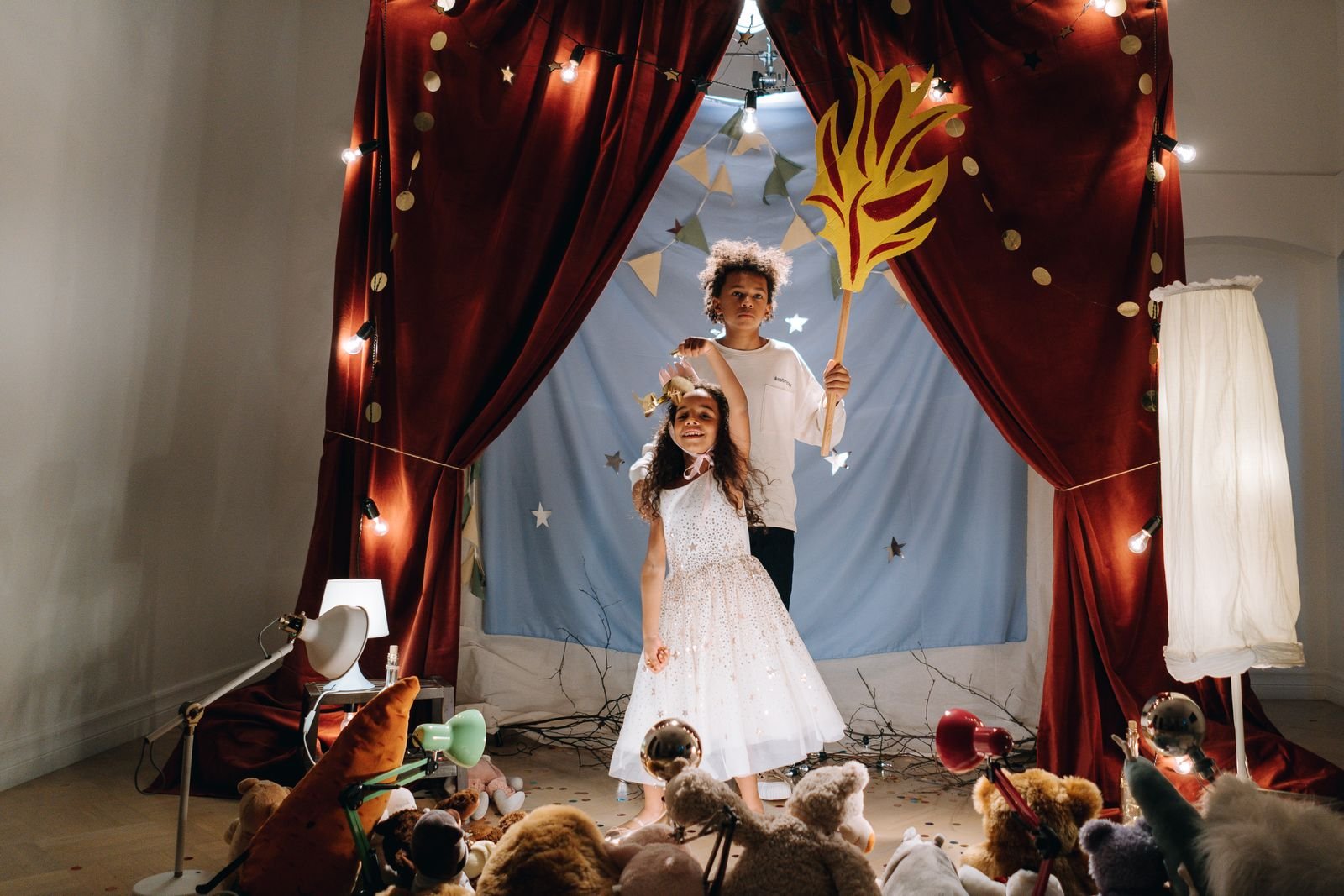 Duas crianças em um palco improvisado com cortinas vermelhas e luzes, em foto ilustrativa de teatro infantil