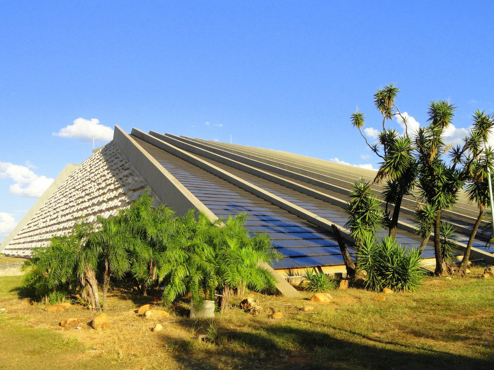 Fachada do Teatro Nacional Cláudio Santoro em Brasília, com a pirâmide de concreto, o relevo lateral e palmeiras em primeiro plano sob céu azul